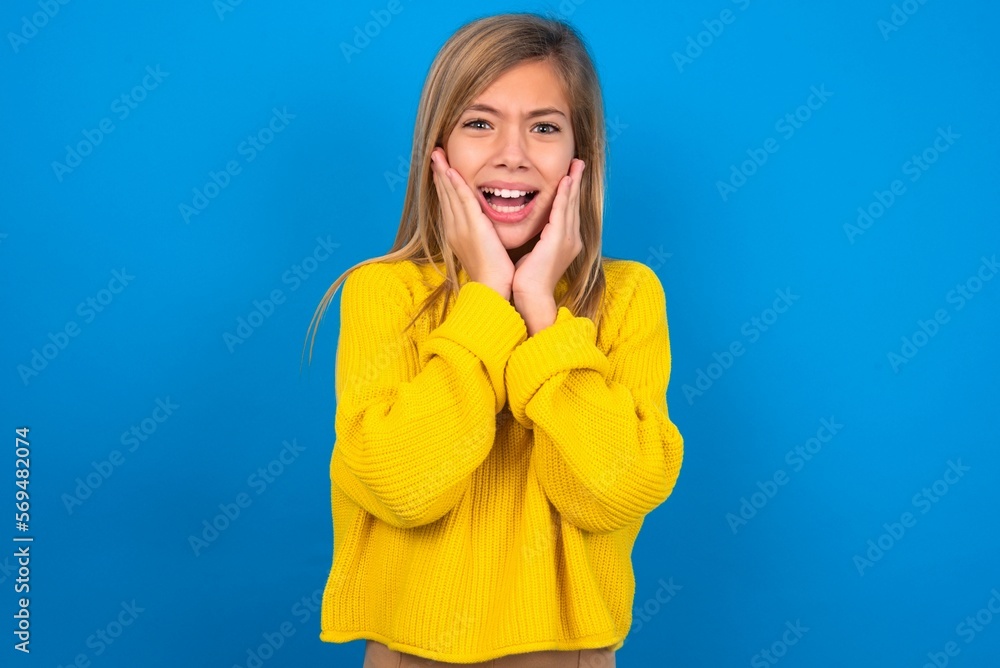 Upset caucasian teen girl wearing yellow sweater over blue studio background touching face with two hands Upset caucasian teen girl wearing yellow sweater over blue studio background touching face with two hands