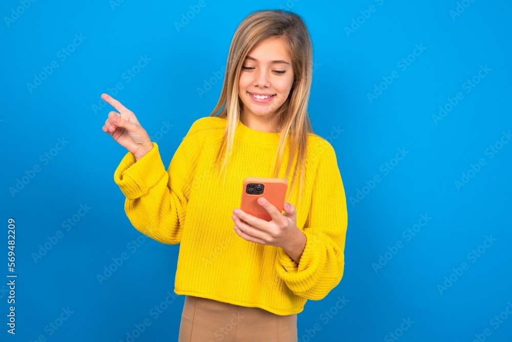 Smiling caucasian teen girl wearing yellow sweater over blue studio background pointing finger at blank space holding phone in one hand Smiling caucasian teen girl wearing yellow sweater over blue studio background pointing finger at blank space holding phone in one hand