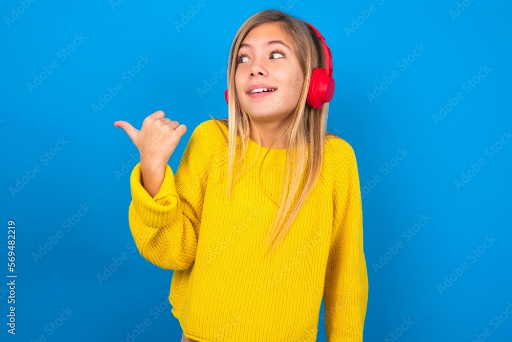 caucasian teen girl wearing yellow sweater over blue studio background listens audio track via wireless headphones points thumb away advertises copy space caucasian teen girl wearing yellow sweater over blue studio background listens audio track via wireless headphones points thumb away advertises copy space