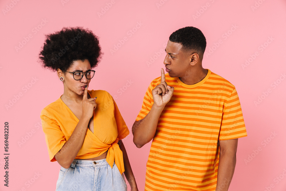 African couple showing silence gesture and looking at each other while standing isolated over pink background African couple showing silence gesture and looking at each other while standing isolated over pink background