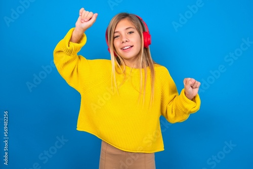 Carefree caucasian teen girl wearing yellow sweater over blue studio background with toothy smile raises arms dances carefree moves with rhythm of music listens music from playlist via headphones