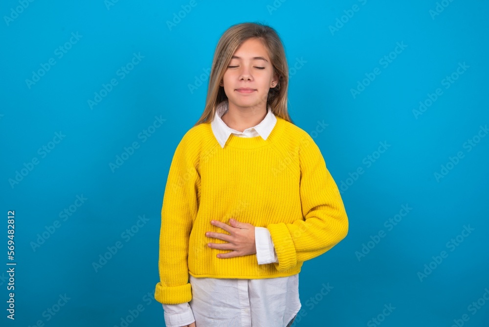 caucasian teen girl wearing yellow sweater over blue studio background touches tummy, smiles gently, eating and satisfaction concept. caucasian teen girl wearing yellow sweater over blue studio background touches tummy, smiles gently, eating and satisfaction concept.