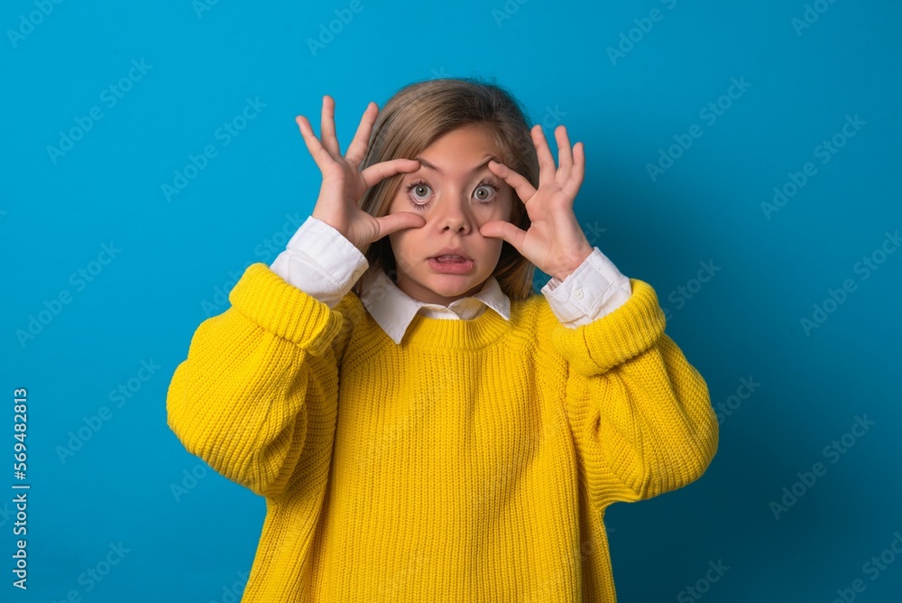 caucasian teen girl wearing yellow sweater over blue studio background keeping eyes opened to find a success opportunity. caucasian teen girl wearing yellow sweater over blue studio background keeping eyes opened to find a success opportunity.