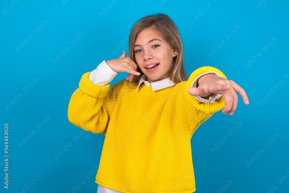 caucasian teen girl wearing yellow sweater over blue studio background smiling cheerfully and pointing to camera while making a call you later gesture, talking on phone