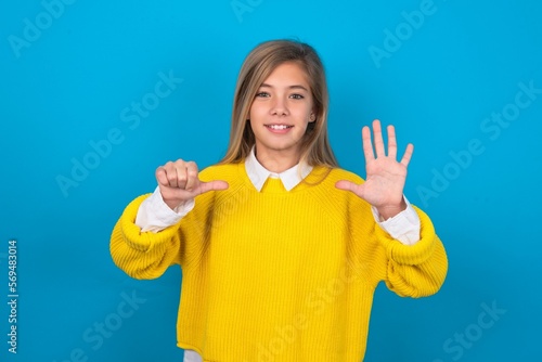 caucasian teen girl wearing yellow sweater over blue studio background showing and pointing up with fingers number six while smiling confident and happy.
