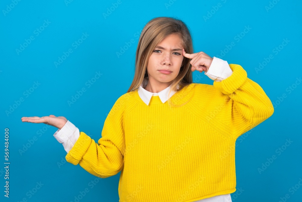 caucasian teen girl wearing yellow sweater over blue studio background, feeling confused and annoyed with open palm showing copy space and pointing finger to forehead. Think about it. caucasian teen girl wearing yellow sweater over blue studio background, feeling confused and annoyed with open palm showing copy space and pointing finger to forehead. Think about it.