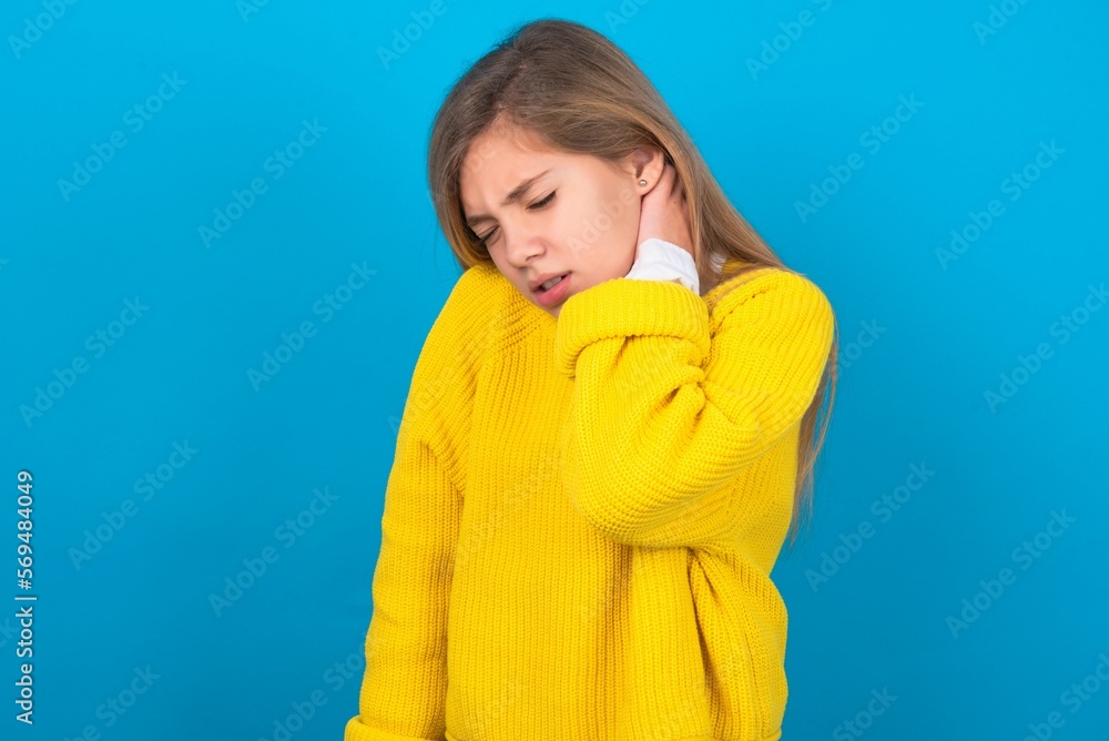 caucasian teen girl wearing yellow sweater over blue studio background Suffering of neck ache injury, touching neck with hand, muscular pain caucasian teen girl wearing yellow sweater over blue studio background Suffering of neck ache injury, touching neck with hand, muscular pain