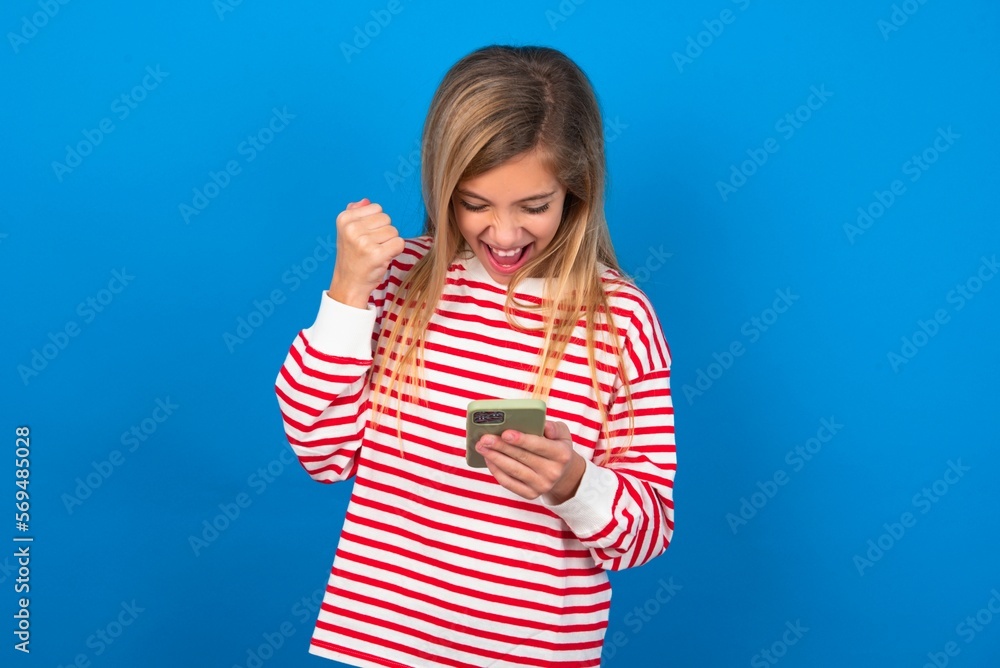 beautiful caucasian teen girl wearing striped T-shirt over blue wall holding in hands cell and rising his fist up being excited after reading good news.