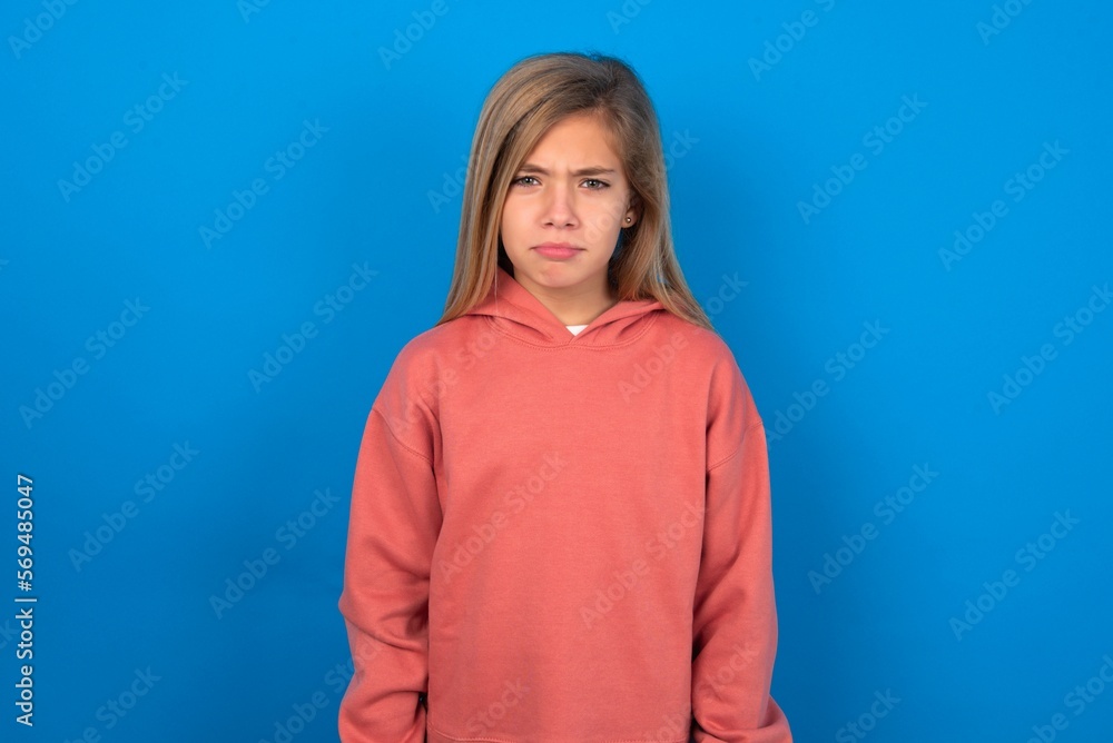 Fototapeta premium Offended dissatisfied beautiful caucasian teen girl wearing pink sweater over blue wall with moody displeased expression at camera being disappointed by something