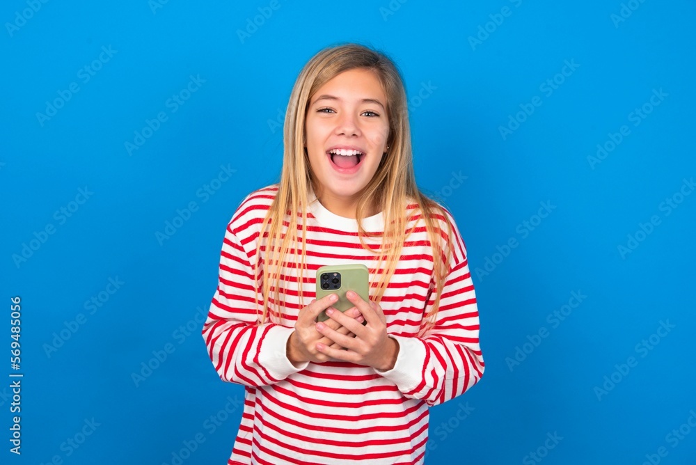 Excited beautiful caucasian teen girl wearing striped T-shirt over blue wall holding smartphone and looking amazed to the camera after receiving good news. Excited beautiful caucasian teen girl wearing striped T-shirt over blue wall holding smartphone and looking amazed to the camera after receiving good news.