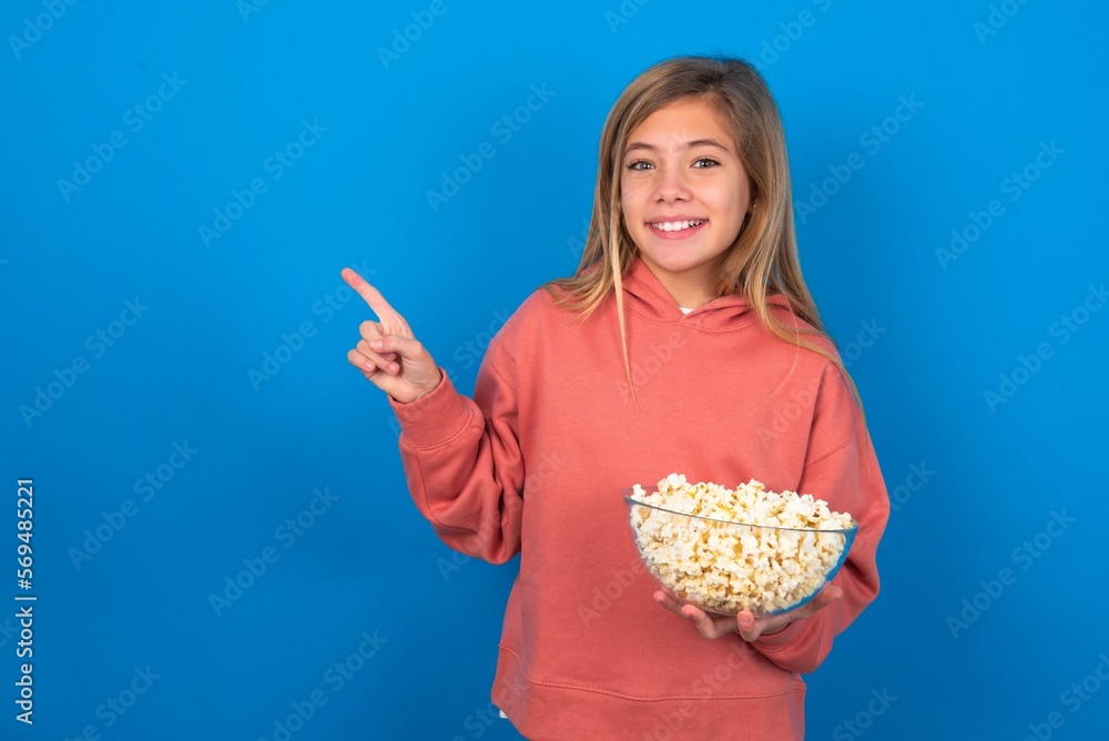 beautiful caucasian teen girl wearing pink sweater over blue wall eating popcorn points at copy space indicates for advertising gives right direction beautiful caucasian teen girl wearing pink sweater over blue wall eating popcorn points at copy space indicates for advertising gives right direction