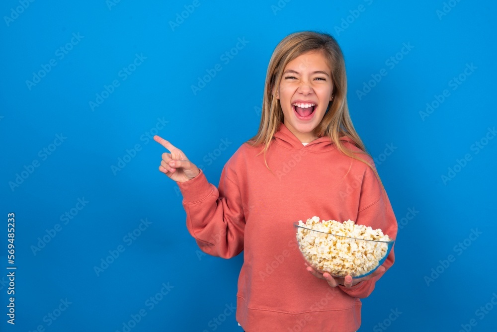 beautiful caucasian teen girl wearing pink sweater over blue wall eating popcorn points aside on copy blank space. People promotion and advertising concept beautiful caucasian teen girl wearing pink sweater over blue wall eating popcorn points aside on copy blank space. People promotion and advertising concept