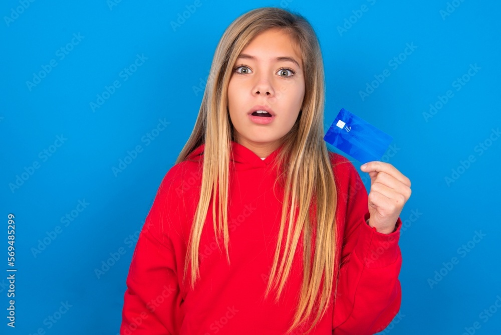 Fototapeta premium Photo of beautiful caucasian teen girl wearing red sweater over blue wall amazed shocked hold credit card payment