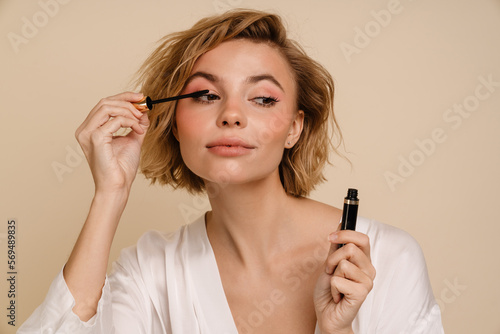 Foto Young woman doing make up with mascara isolated over beige wall