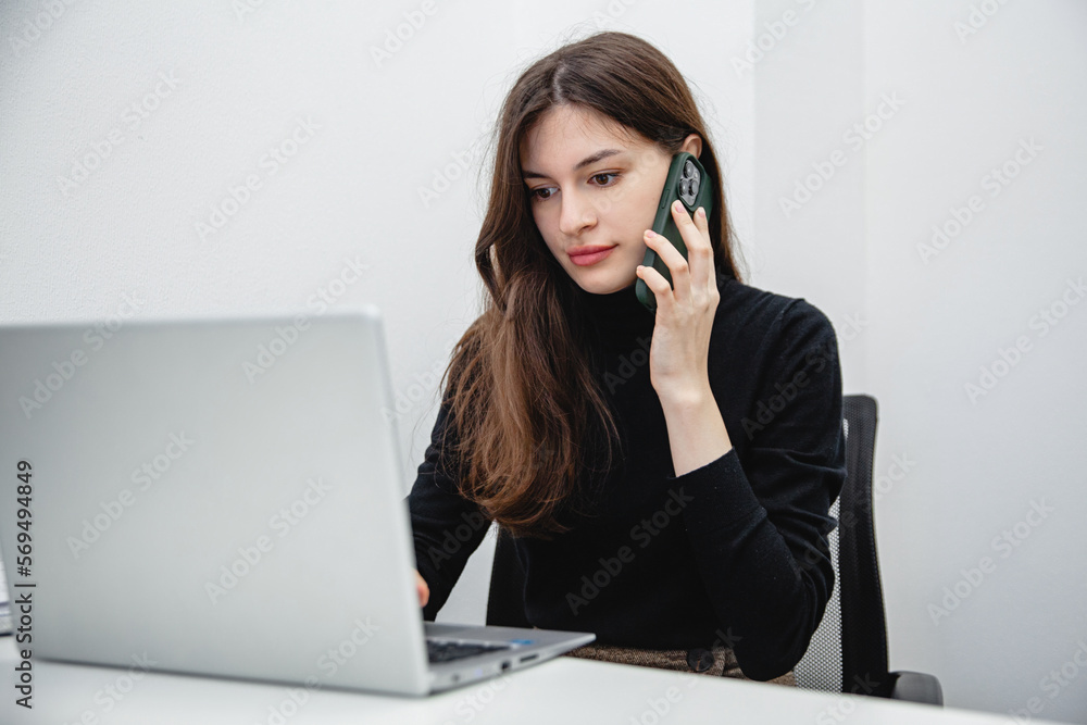 A young girl with long dark hair in black clothes sitting at a white table in the office and talks on the phone looking into a white laptop A young girl with long dark hair in black clothes sitting at a white table in the office and talks on the phone looking into a white laptop