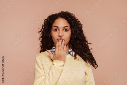 Canvas-taulu Studio portrait of a young multiracial surprised beautiful woman with curly hair covering her mouth with hand, isolated over beige background