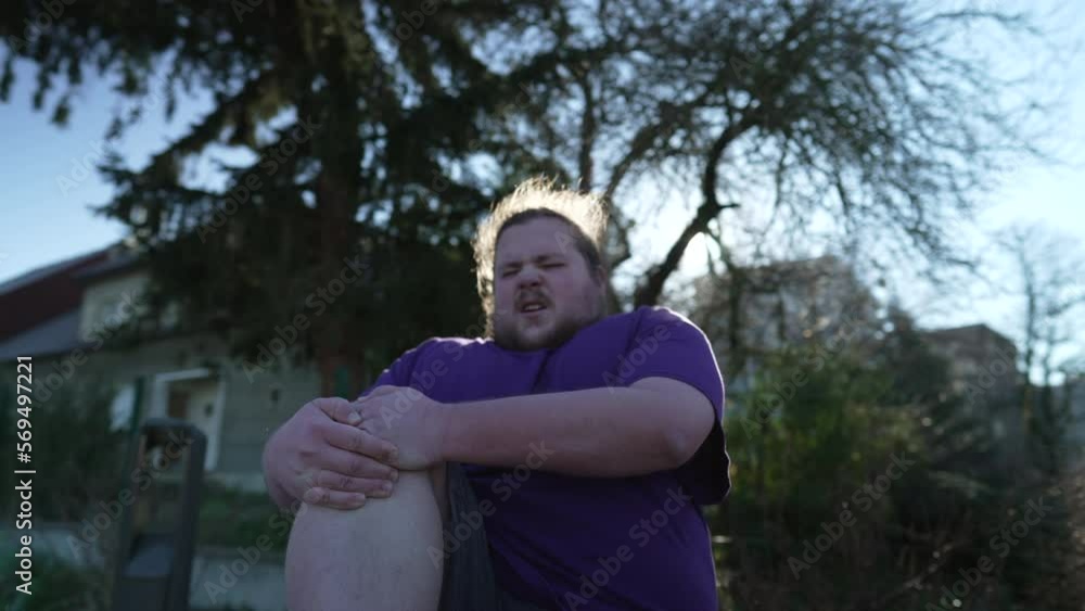 Person suffering from knee pain sitting at bench outdoors. One overweight caucasian male person having physical injury Person suffering from knee pain sitting at bench outdoors. One overweight caucasian male person having physical injury