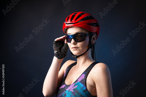 A young female cyclist wearing a safety helmet and glasses, dressed in a bib shorts poses against a black background in the studio. © Budjak Studio