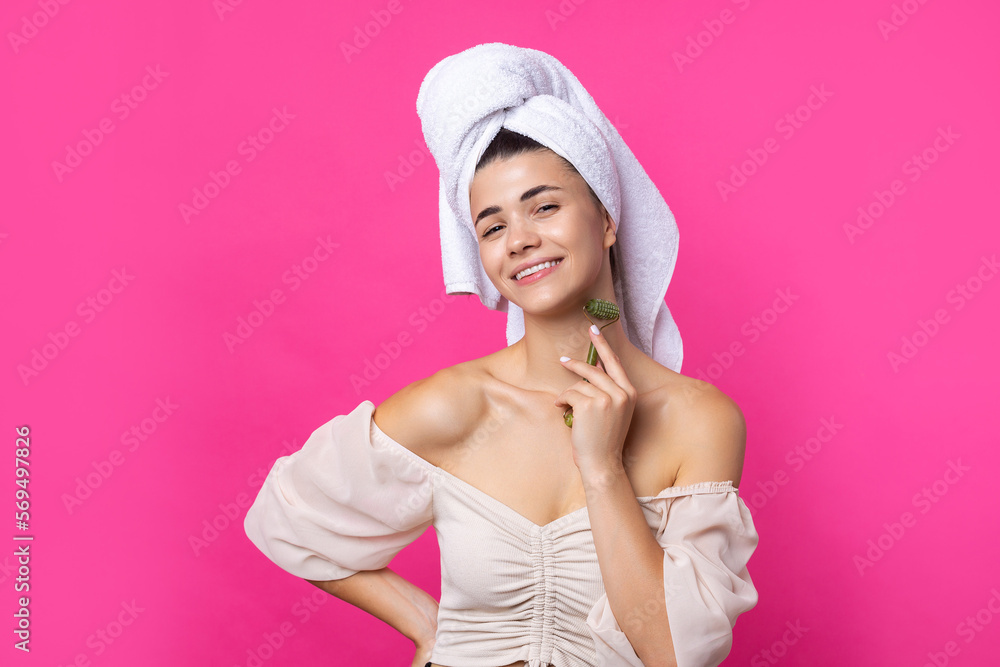 A beautiful cheerful attractive girl with a towel on her head holds a cosmetic roller near her face against a pink background. A beautiful cheerful attractive girl with a towel on her head holds a cosmetic roller near her face against a pink background.