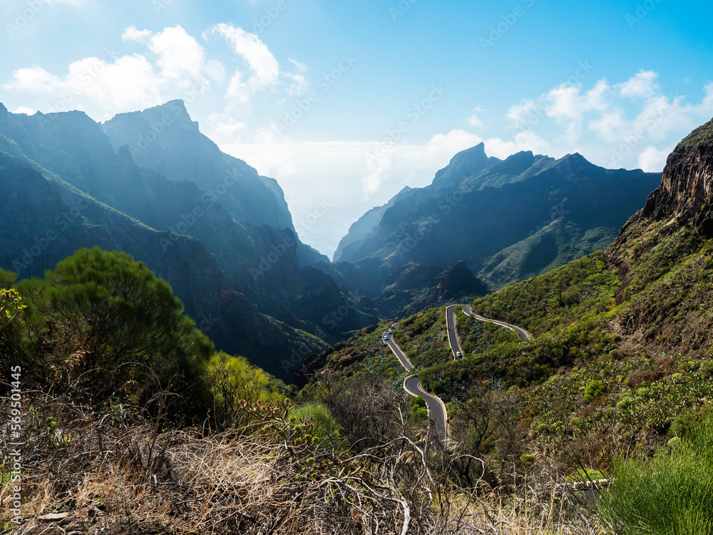 Fototapeta premium Picturesque landscape with winding curvy mountain road towards Masca village. Green valley, ocean, massive cliffs and sharp rock formation. Tenerife, Canary Islands, Spain. sunny winter day, blue sky