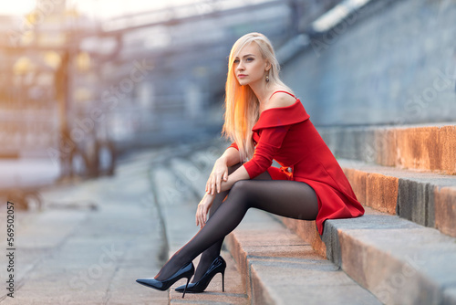 Fototapeta Beautiful blonde girl in a red dress with perfect legs in pantyhose and shoes with high heels posing outdoor on the city square