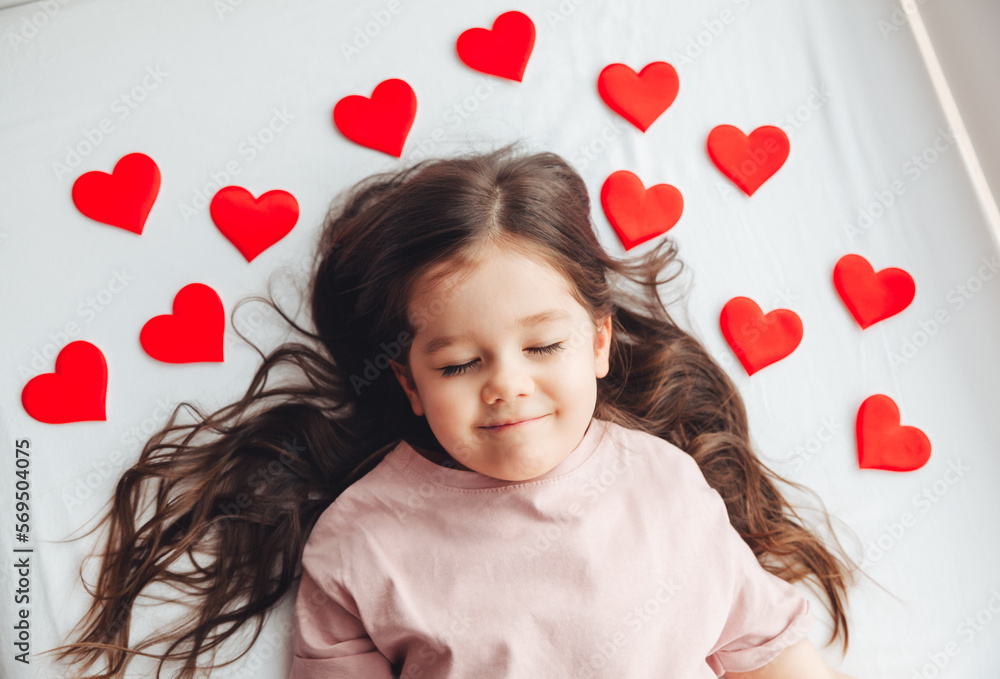 Valentine's Day, a little girl lying at home on a white bed among hearts, smiling and laughing with happiness, congratulating on the holiday Valentine's Day, a little girl lying at home on a white bed among hearts, smiling and laughing with happiness, congratulating on the holiday
