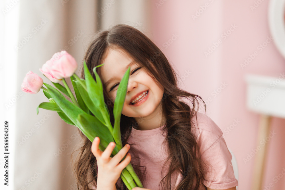 Naklejka premium Portrait of a little girl with long dark hair close-up. The baby hugs a bouquet of fresh, delicate pink tulips. A gift for the holiday, spring time.