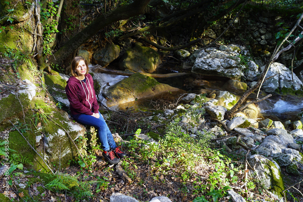 Naklejka premium A smiling girl is resting on a rock near a picturesque forest stream during a hike