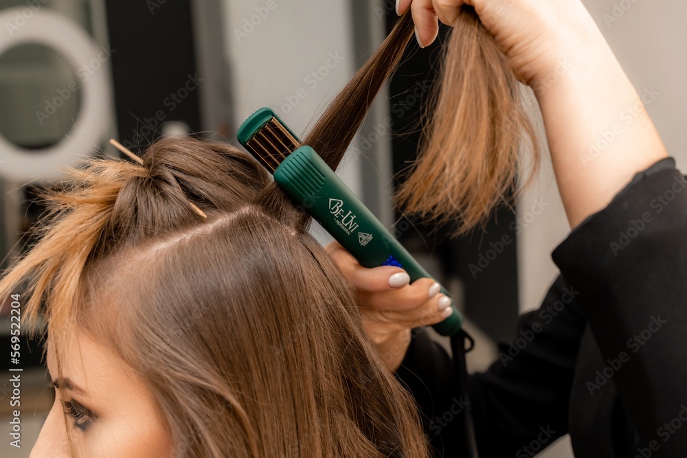 Naklejka premium The hairdresser prepares a woman, makes a hairstyle with curls using a curling iron. Long light brown natural hair. Self care concept.