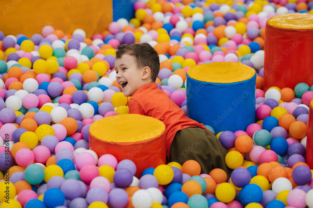 Obraz premium A happy little boy has fun jumping into a dry pool with colorful balls. The kid boy is playing and having a good time in the game room with colorful balls.
