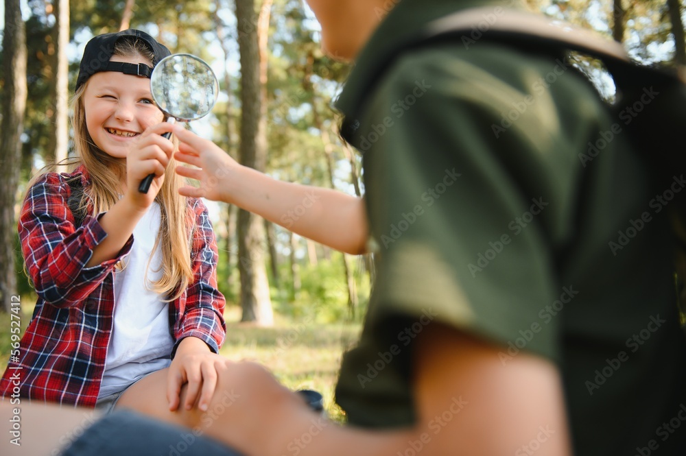 Group of curious happy school kids in casual clothes with backpacks exploring nature and forest together on sunny autumn day, girl holding magnifying glass and looking at fir cone in boy hands.