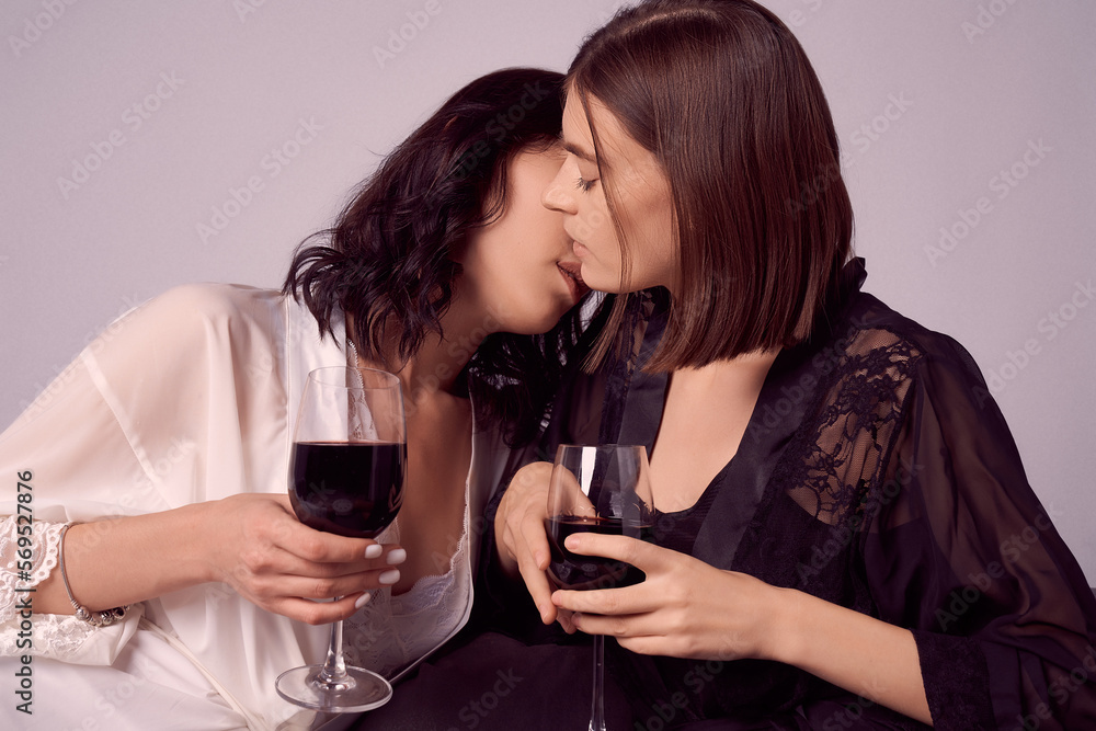 Valentine's day concept. Portrait of two lesbians in satin negligee holding glasses of red wine while sitting on a sofa, intimate moments of private life. Two girls are relaxing on a couch at home. Valentine's day concept. Portrait of two lesbians in satin negligee holding glasses of red wine while sitting on a sofa, intimate moments of private life. Two girls are relaxing on a couch at home.