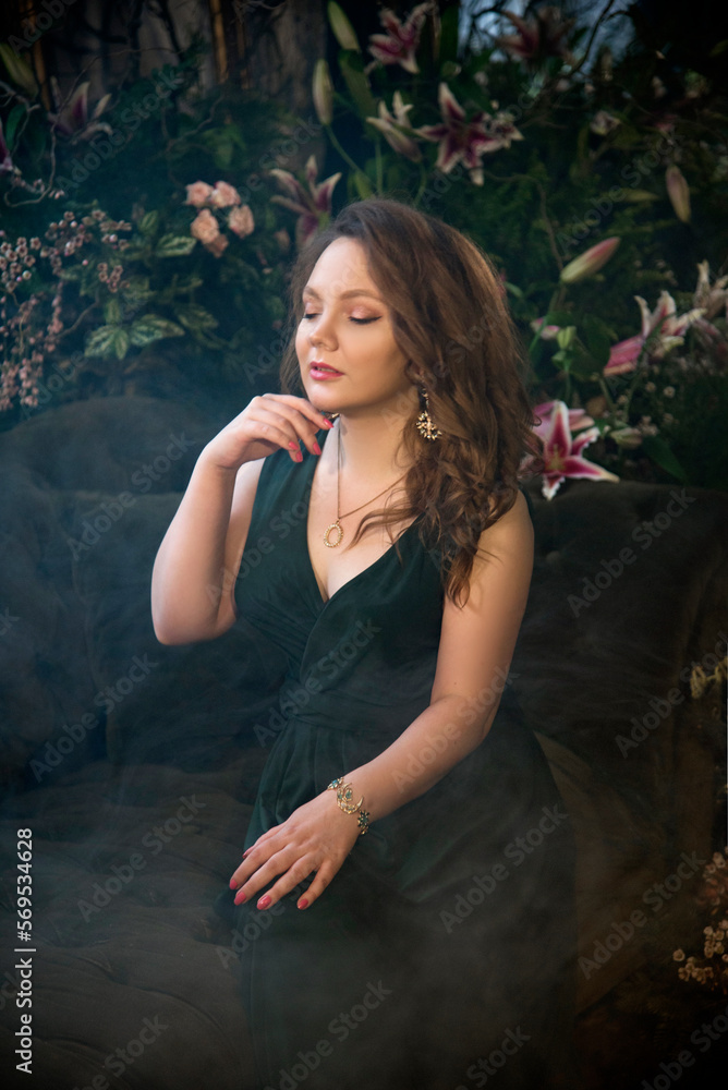 Fototapeta premium portrait of a beautiful brunette smiling woman in vintage room with flowers and smoke in dark green long dress