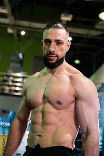 A brutal muscular man with a beard and a perfect body looks at the camera in the gym. Fitness, beauty