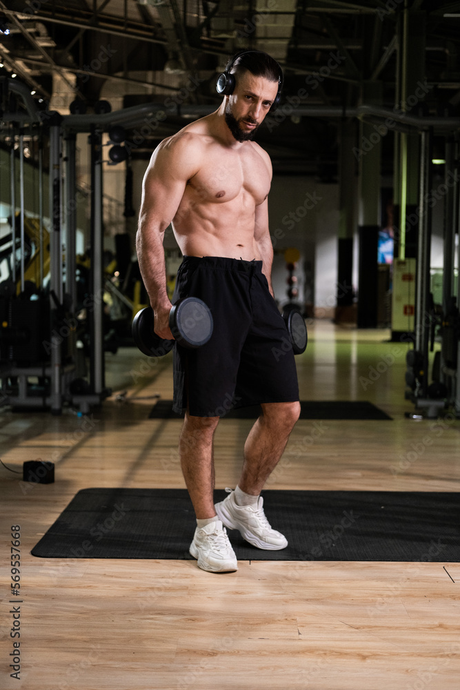 Fototapeta premium A brutal muscular man with a beard and a perfect body wearing headphones does exercises with dumbbells in the gym. Fitness, beauty