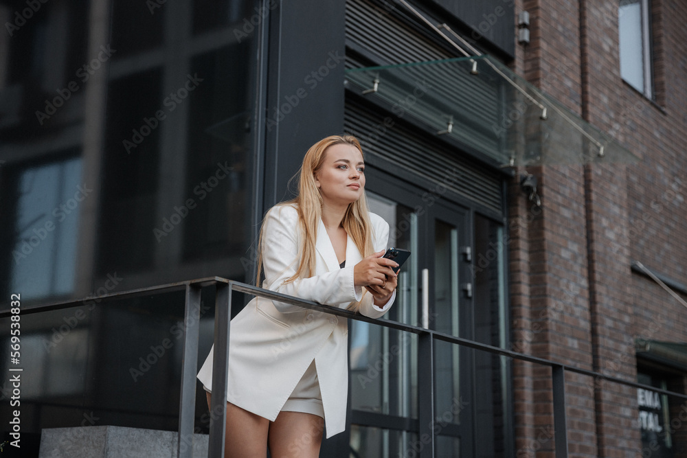 Blond stylish woman at oversized white jacket and phone on hand posing at the railing and steps on the city background Blond stylish woman at oversized white jacket and phone on hand posing at the railing and steps on the city background