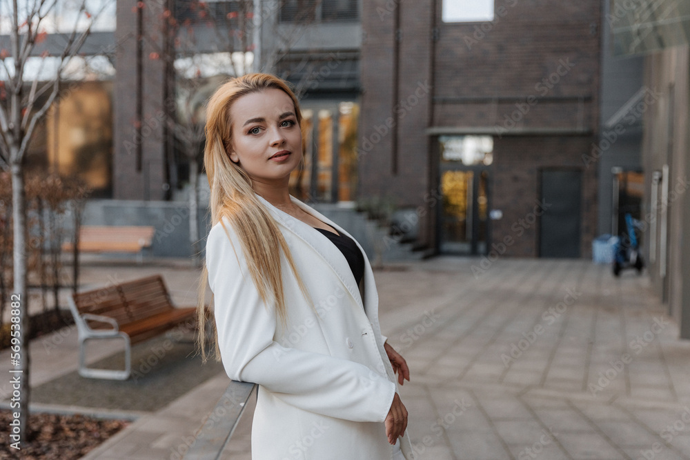 Blond stylish woman at oversized white jacket posing at the railing and steps on the city background