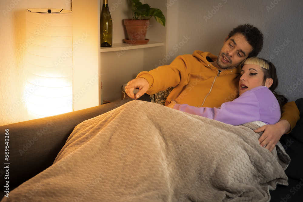 Biracial couple stretched out on the sofa covered with the blanket watching and commenting on their favorite series on TV Biracial couple stretched out on the sofa covered with the blanket watching and commenting on their favorite series on TV