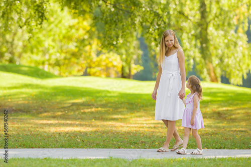 Little daughter and young adult mother in dresses speaking and walking on sidewalk at city park. Spending time together in beautiful warm sunny summer day. Side view.