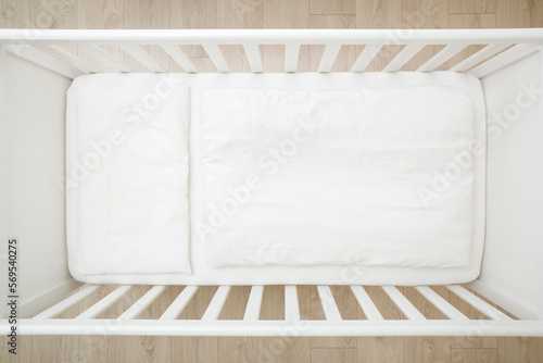 Empty white baby crib with mattress, sheet, pillow and blanket. Closeup. Top down view.