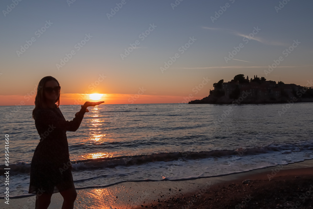 Silhouette of barefoot woman on sand beach with scenic romantic view on idyllic island Sveti Stefan at sunset, Budva Riviera, Adriatic Mediterranean Sea, Montenegro, Europe. Summer vacation at seaside