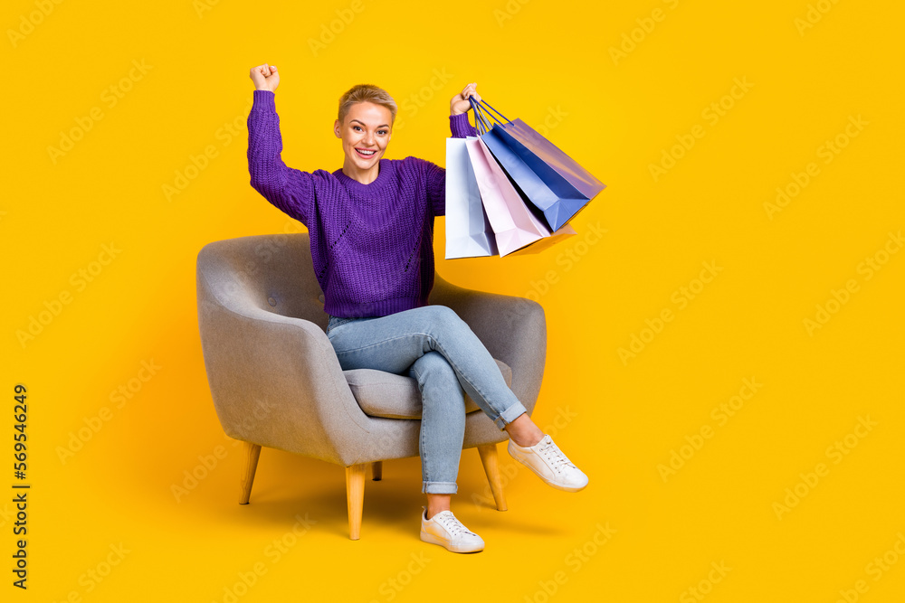 Full body portrait of crazy delighted lady sit chair hold bags raise fist celebrate luck isolated on yellow color background Full body portrait of crazy delighted lady sit chair hold bags raise fist celebrate luck isolated on yellow color background