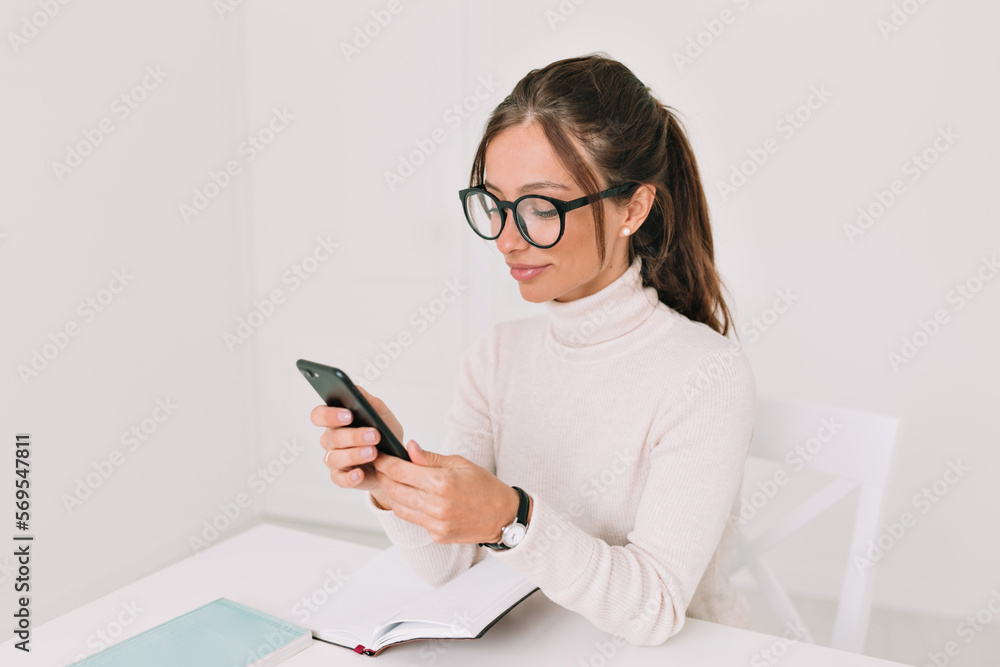 Lovely cute woman in pullover and glasses is cheerfully talking on phone and sitting at table with laptop at home stylish office. Indoor photo of enthusiastic European woman chatting online in phone
