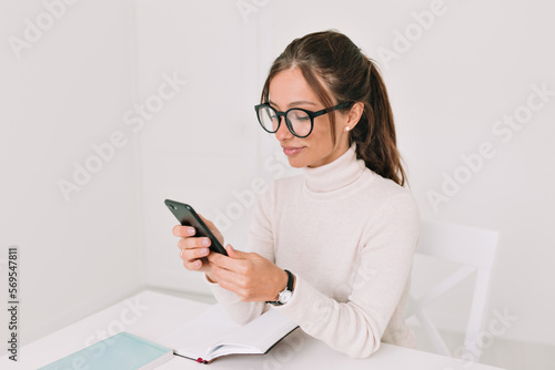 Obraz na płótnie Lovely cute woman in pullover and glasses is cheerfully talking on phone and sitting at table with laptop at home stylish office. Indoor photo of enthusiastic European woman chatting online in phone w salonie