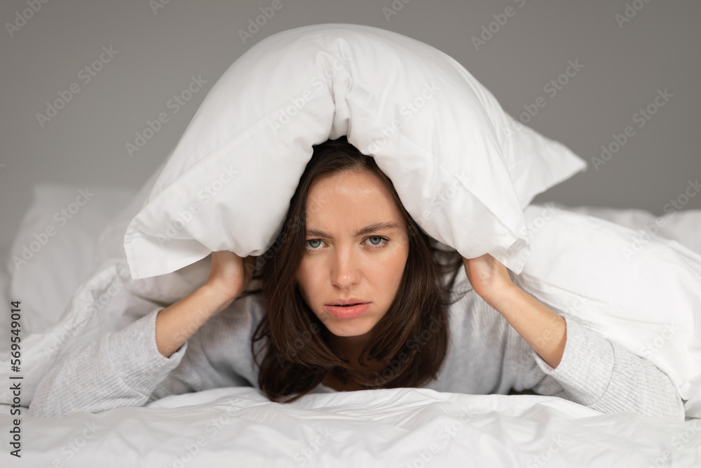 Tired sad caucasian millennial female lies on bed under white blanket, covers head with pillow, suffers from insomnia Tired sad caucasian millennial female lies on bed under white blanket, covers head with pillow, suffers from insomnia