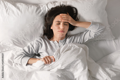 Unhappy sad tired caucasian millennial female lies on bed, suffers from headache, migraine in bedroom photo