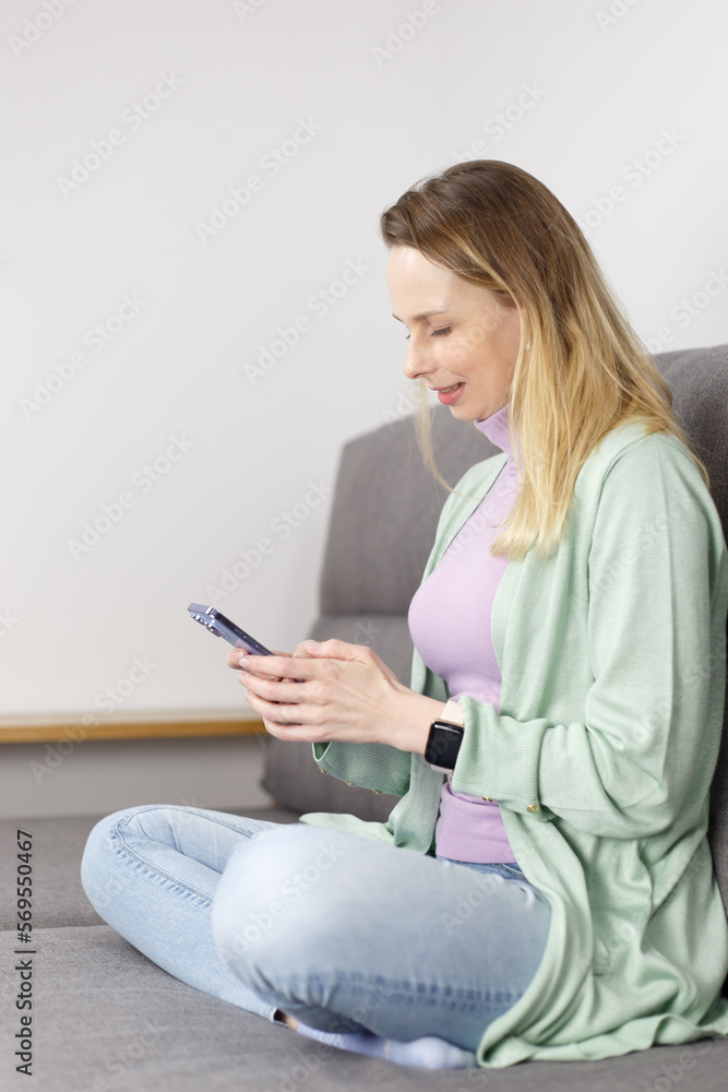 Cheerful adult woman sitting on couch at home and typing sms message on smart phone. White female in early 40s using modern mobile phone for communication Cheerful adult woman sitting on couch at home and typing sms message on smart phone. White female in early 40s using modern mobile phone for communication