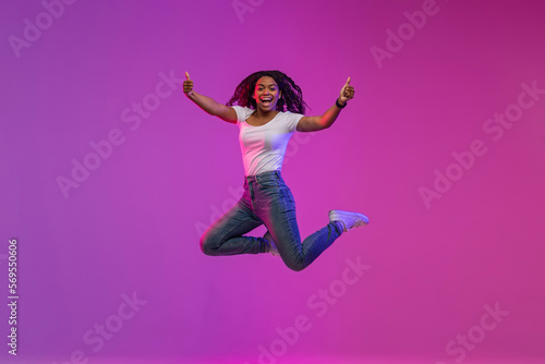 Cheerful Black Woman Jumping In Neon Light And Showing Thumbs Up