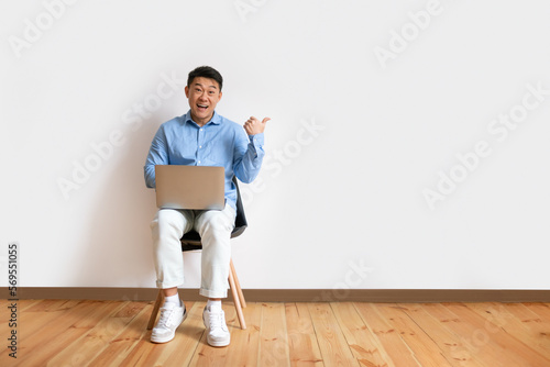 Cheerful middle aged korean man sitting on chair with laptop, pointing aside at empty space, white wall, full length