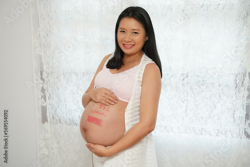 Happy pregnant beautiful Asian woman holding belly with painting loading 99 percent.  touching at belly with smile stand near window at home. prenatal, pregnancy, motherhood concept